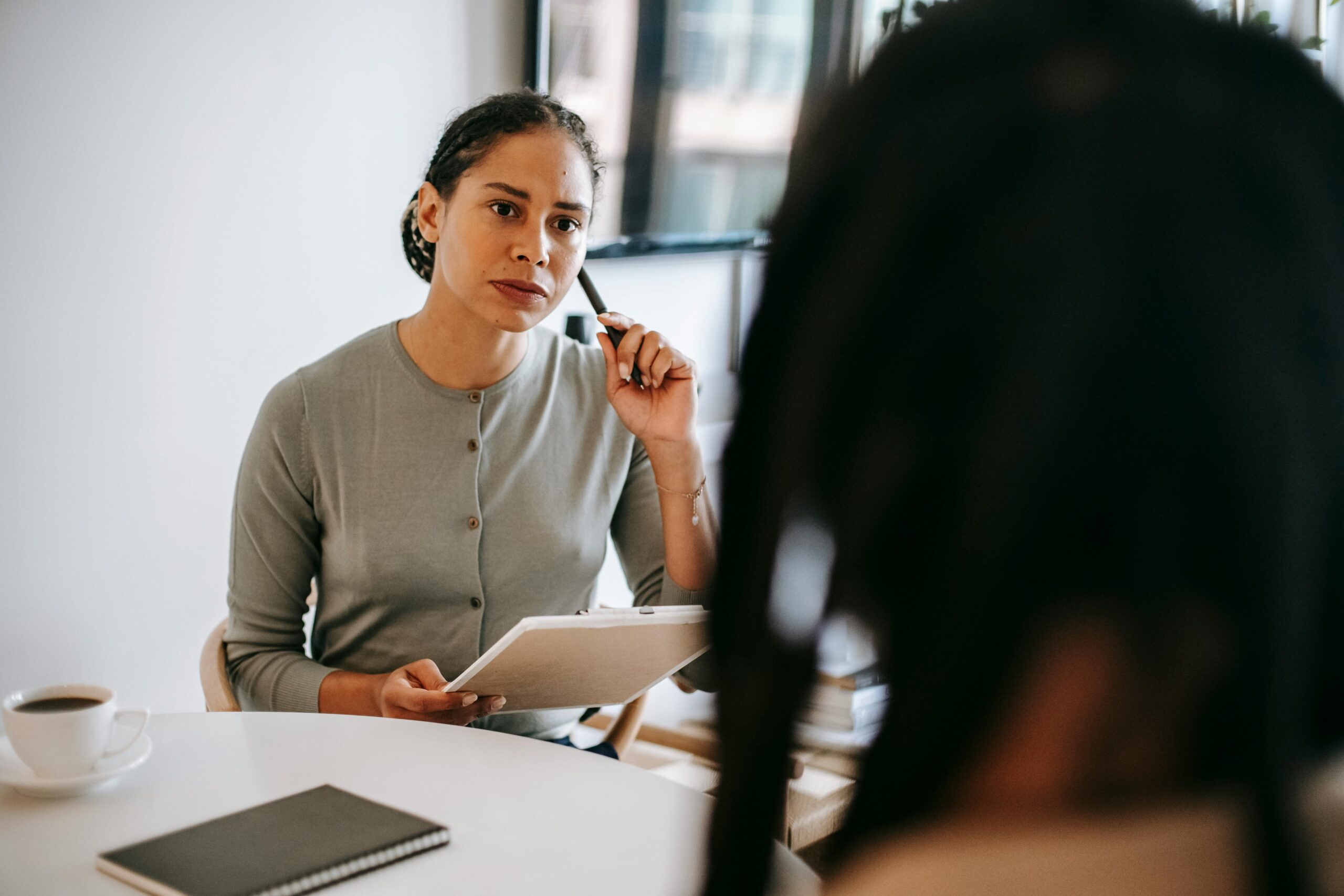 Focused ethnic female counselor in formal wear with clipboard and pen listening to patient problems during psychotherapy session